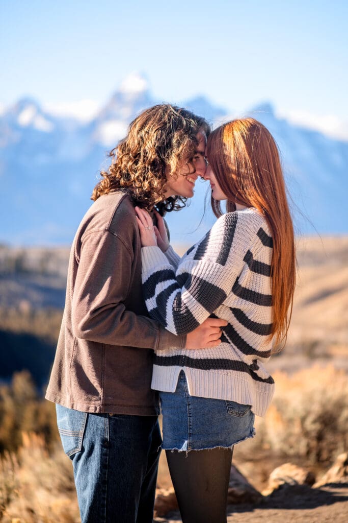 Couple facing each other and smiling with her hands on his chest and a view of the Grand Tetons behind them.