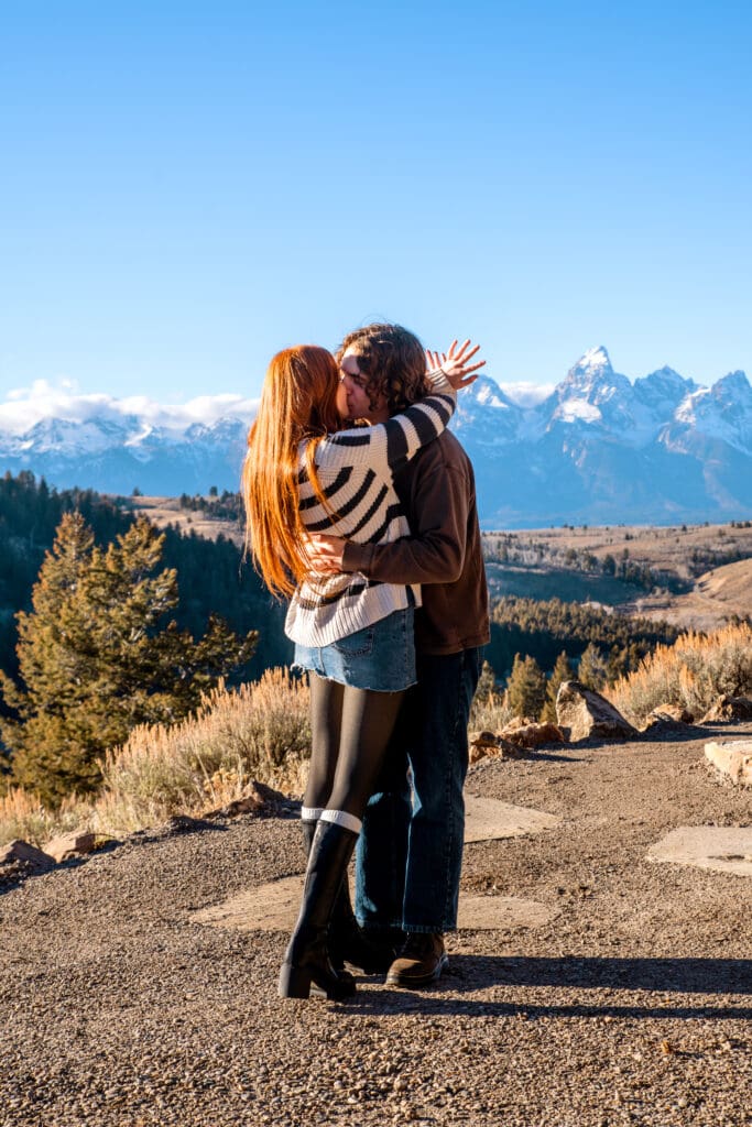 Woman kissing man and showing off engagement ring in front of view of the Grand Tetons.