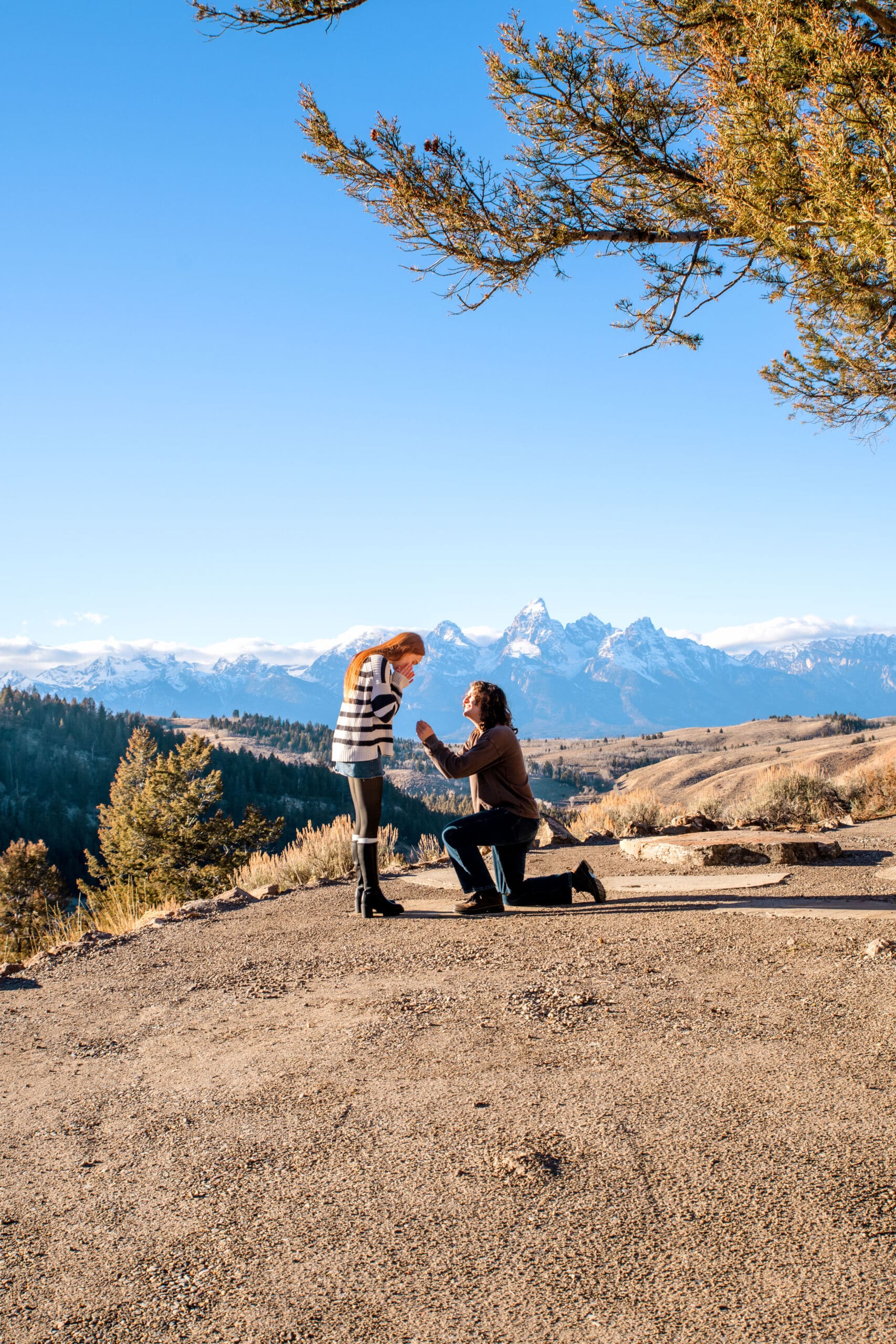 Woman surprised as man proposes in front of view of the Grand Tetons.