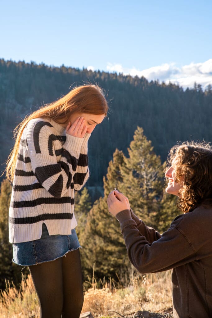 Woman surprised as man proposes in front of view of pine tree hill.