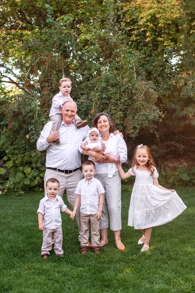 Grandparents pose with five grandkids, with one sitting on grandpa's shoulders, one being held by grandma, one flaring a dress, and two more holding hands.