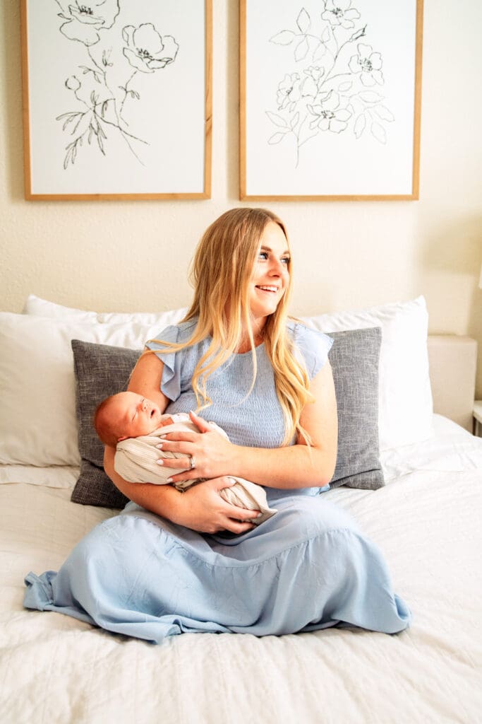 Mother sitting on bed in light blue dress, laughing and looking out the window while holding newborn.