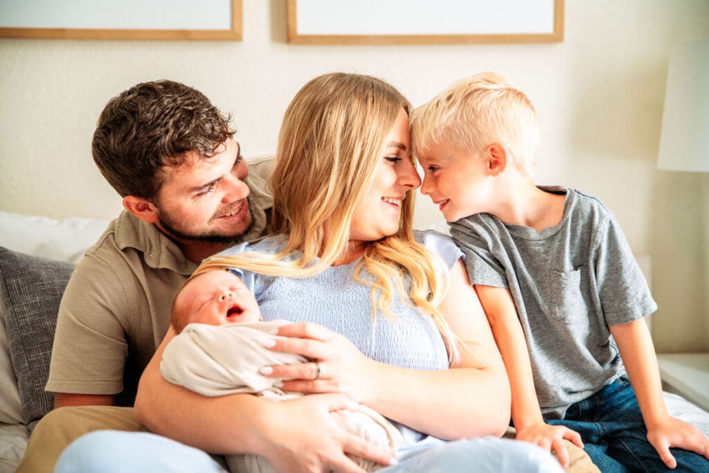 Family sitting on bed with father wrapped around mother, mother holding newborn, and young boy touching forehead with mother's forehead.
