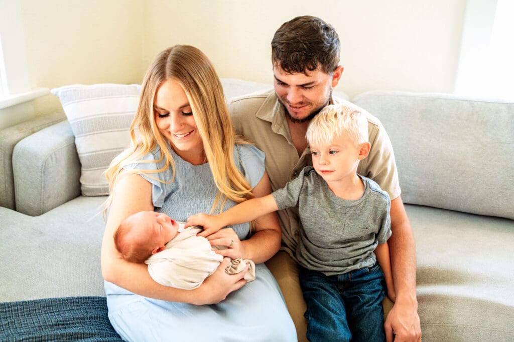 Family sitting on couch and smiling at newborn with father wrapped around mother and young boy, mother holding newborn, and young boy pointing at newborn.