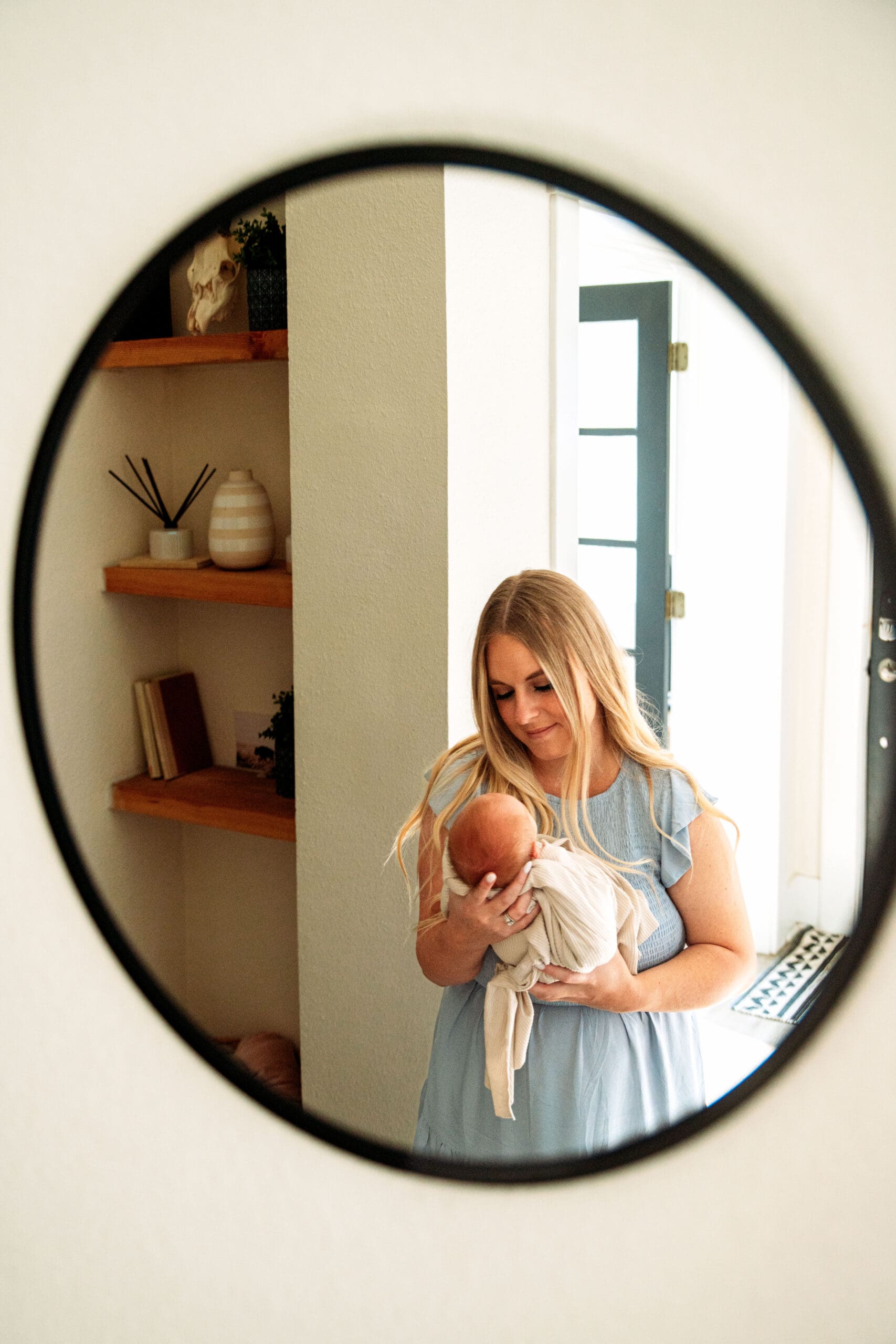 Mother smiling down at newborn, standing in home hallway, seen from a mirror refelction/