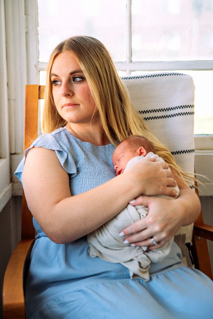 Mother in light blue dress holds newborn and looks over her shoulder out the window while sitting in a rocking chair in an enclosed porch.