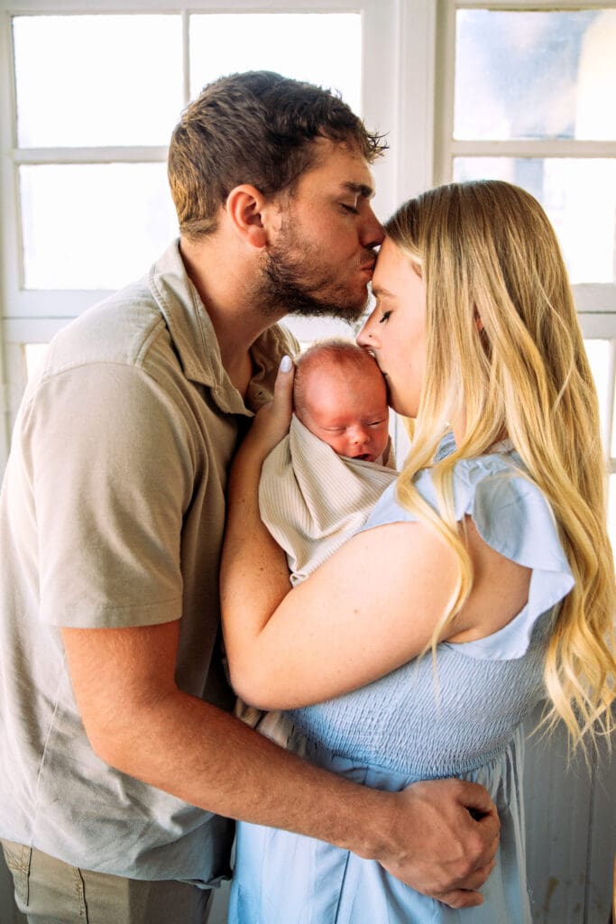 Family standing on enclosed porch with father facing, holding, and kissing mother, and mother facing father and newborn and kissing newborn.