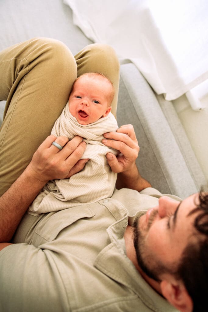 Father tickling newborn who smiles while laid on father's legs.