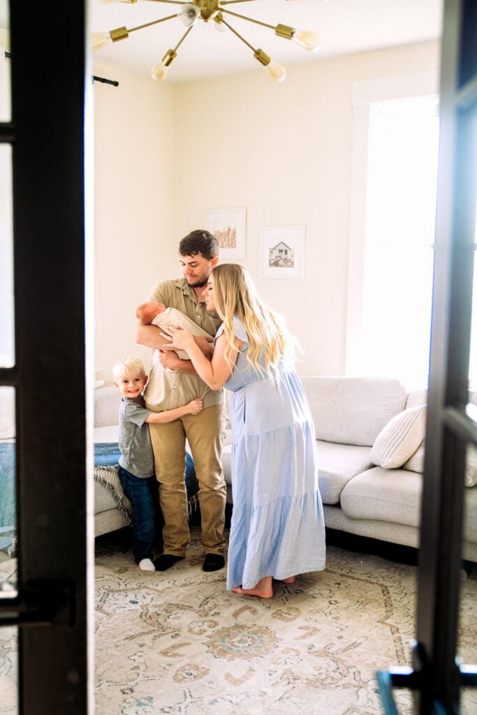 Family standing in living room with father holding newborn, mother tickling newborn, and younger brother smiling at the camera.