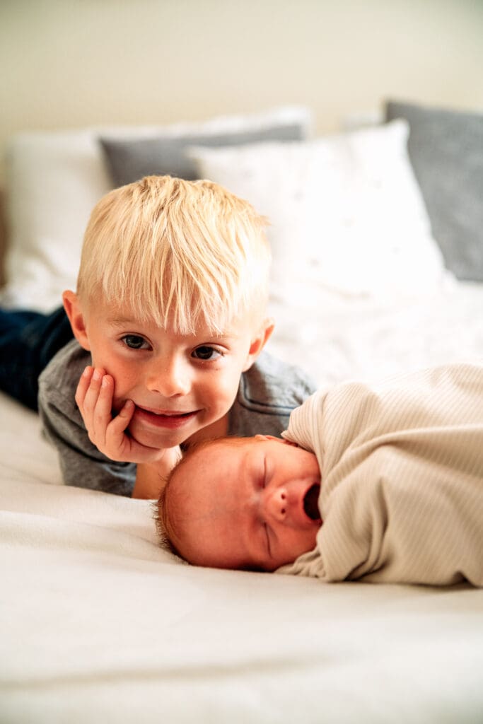 Young boy smiling at camera with his head propped on his head and his newborn sister on the bed with him.