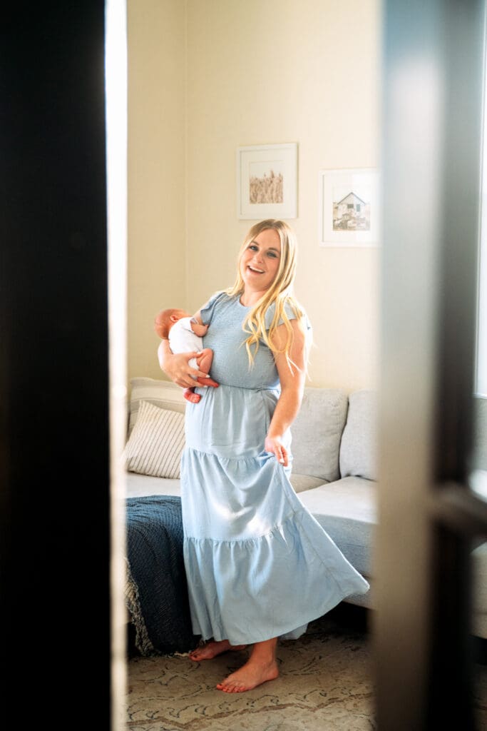 Mother looking at the camera, swaying in light blue dress with a newborn in her arms in a living room, framed by entry doors.