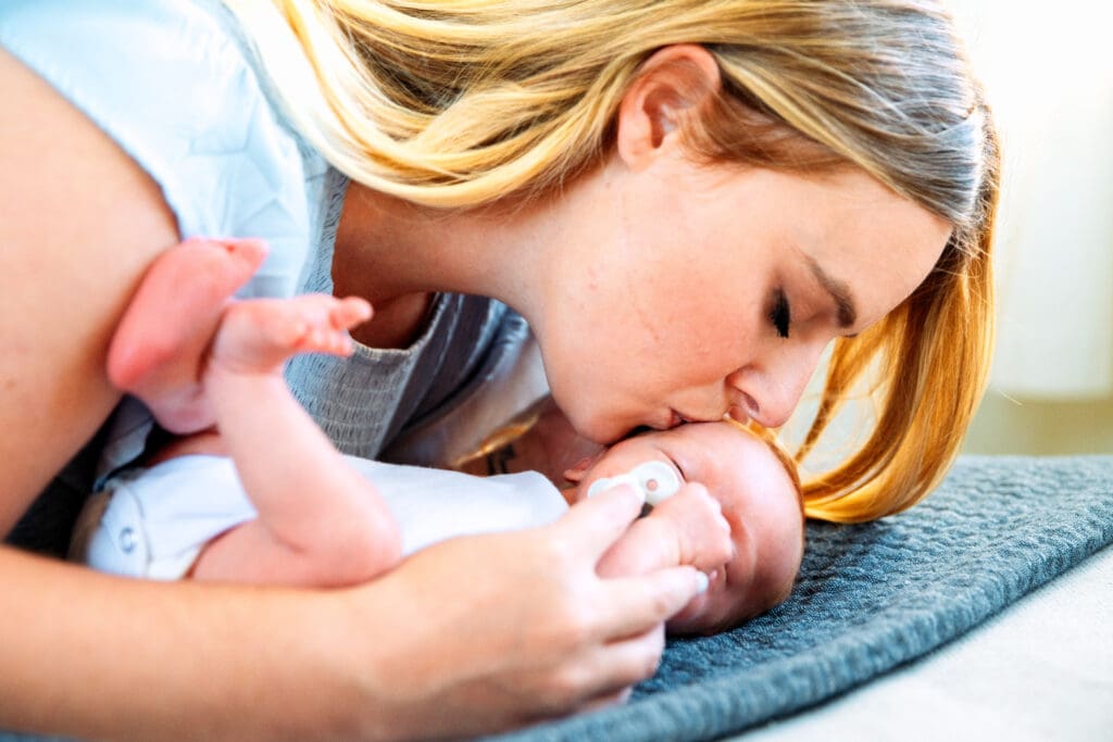 Newborn laying on couch with mother leaning over and kissing newborn's cheek.