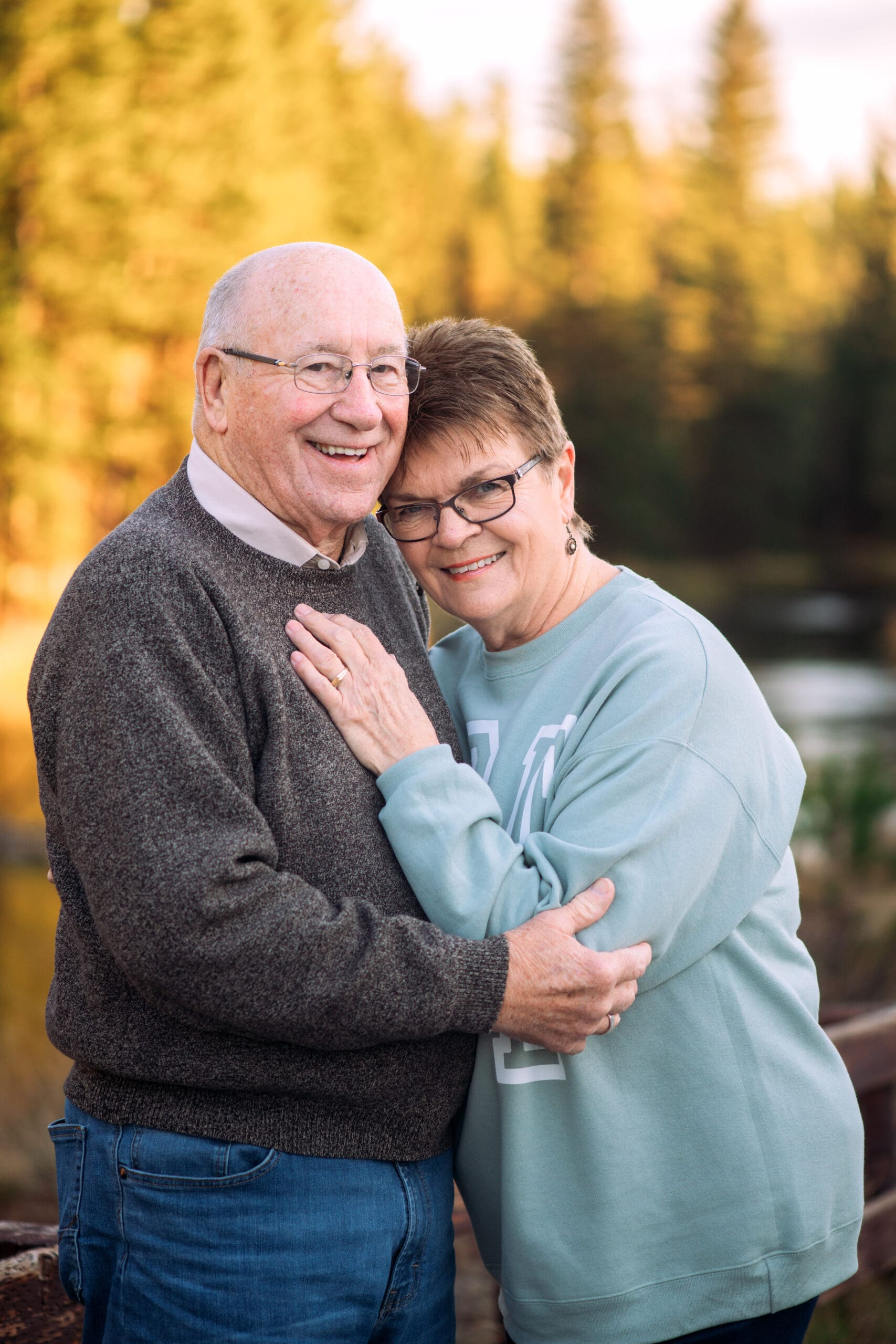 Grandpa holding grandma while grandma leans on him and has her hand on his chest in front of riverwalk.