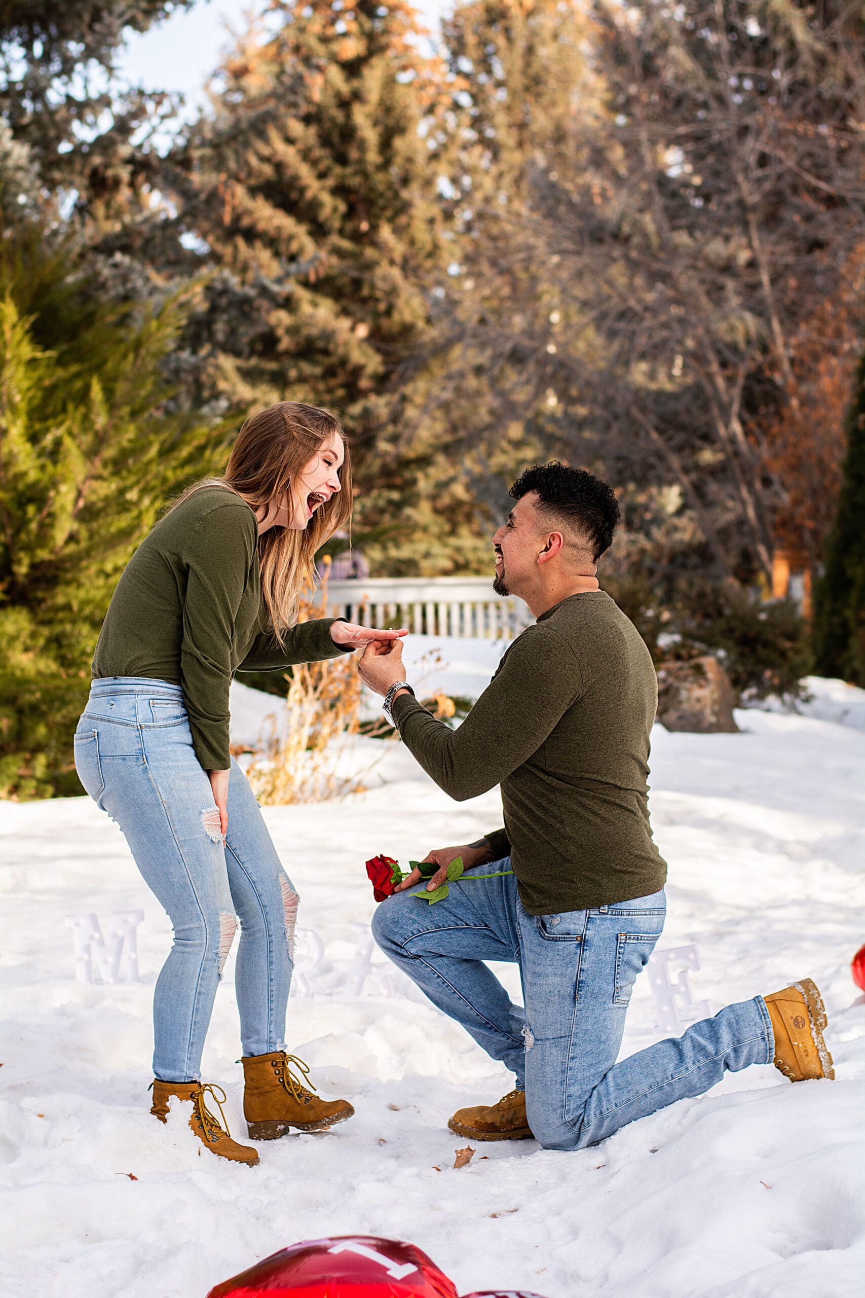 Man down on one knee proposing while woman smiles happily and accepts ring on finger, with red heart balloons on ground in Idaho wintery landscape.