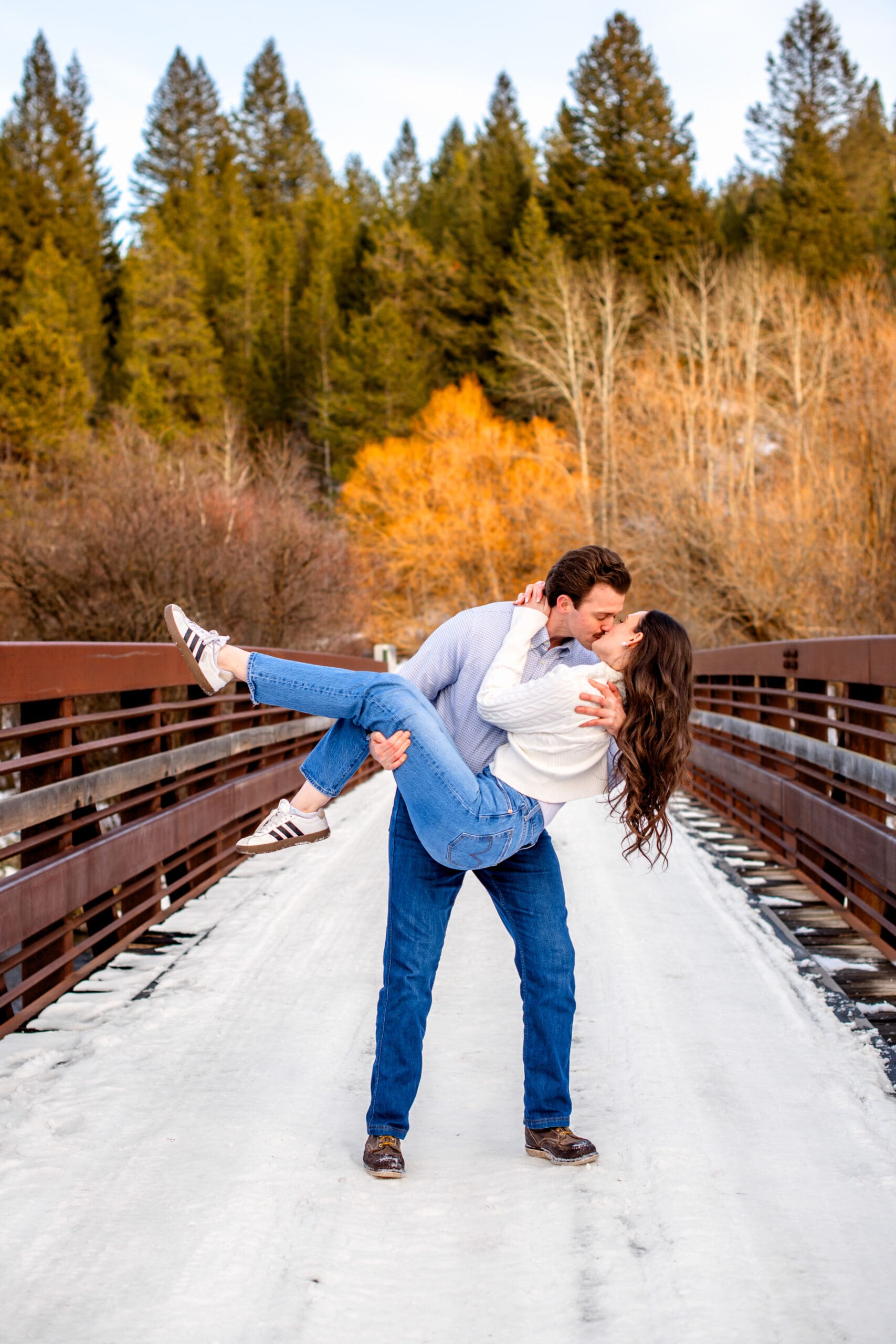 Man carrying and dip kissing woman as woman wraps her arms around man's neck and kicks her legs, on snow covered bridge with green, orange, and bare trees in the background.
