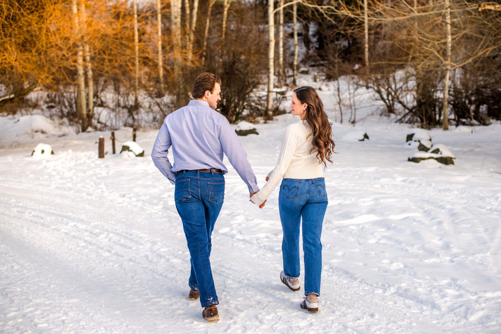 Man and woman holding hands and smiling at each other as they walk away from camera in snowy landscape.