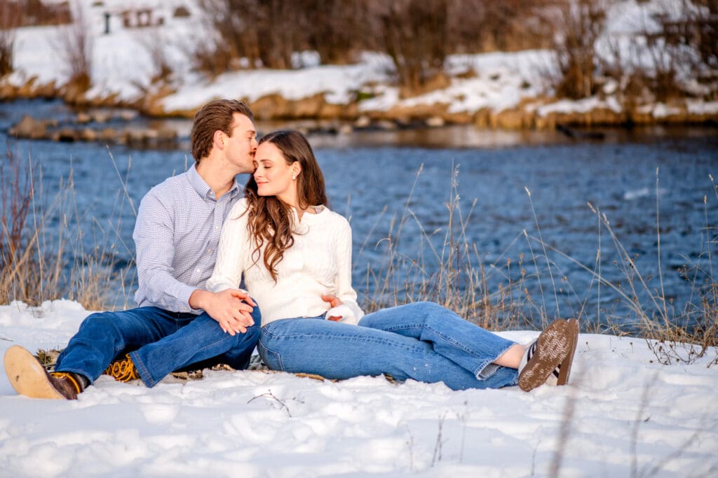 Man and woman sitting in snow and holding hands with man kissing woman's forehead and woman smiling, with river in the background.