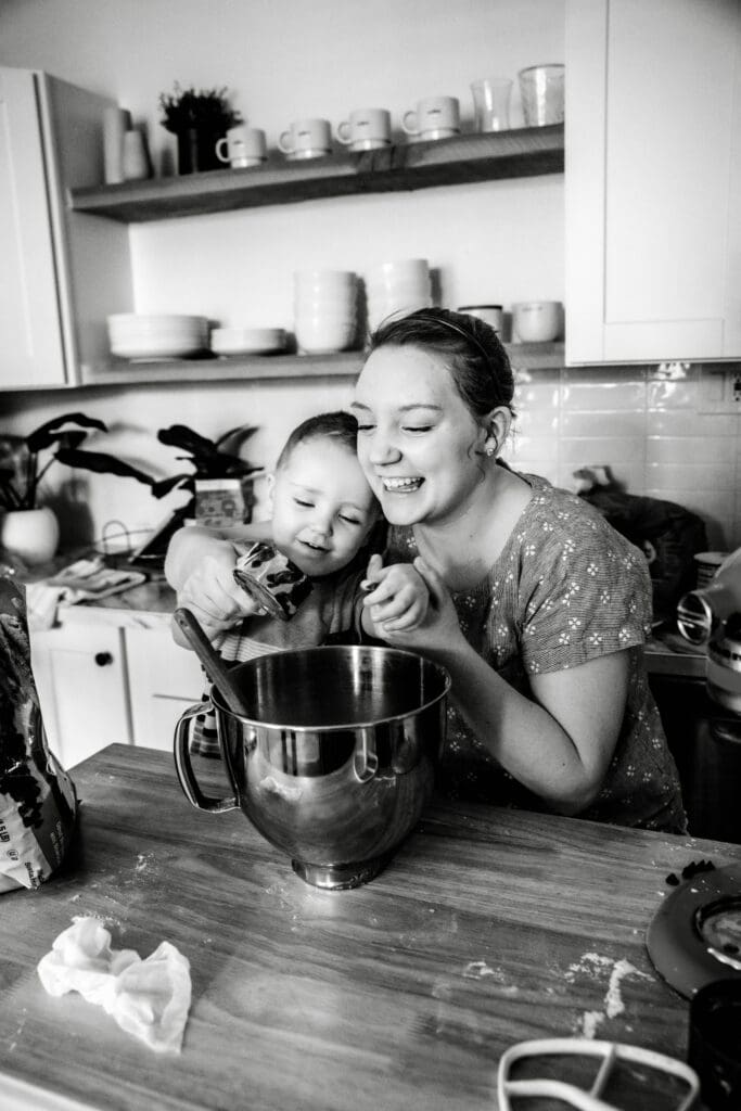 Mom and son smiling as mom helps the young boy add ingredients to mixing bowl on top of messy kitchen counter.