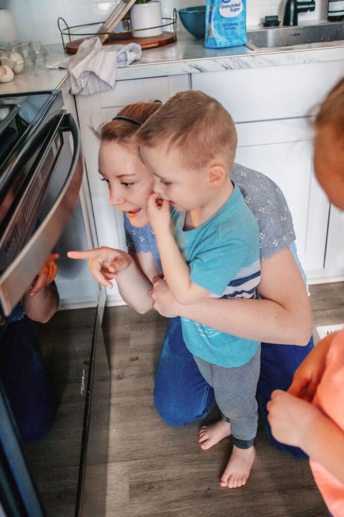 Mom kneeling on kitchen ground in kitchen and holding young boy to her side as she points to oven and explains the cookies baking.