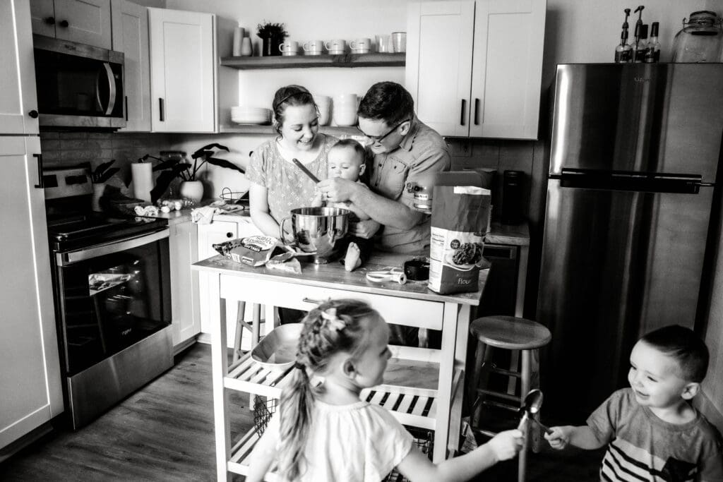 Parents stirring mixing bowl with toddler at island in kitchen while two young children sword fight with spoons in front in black and white.