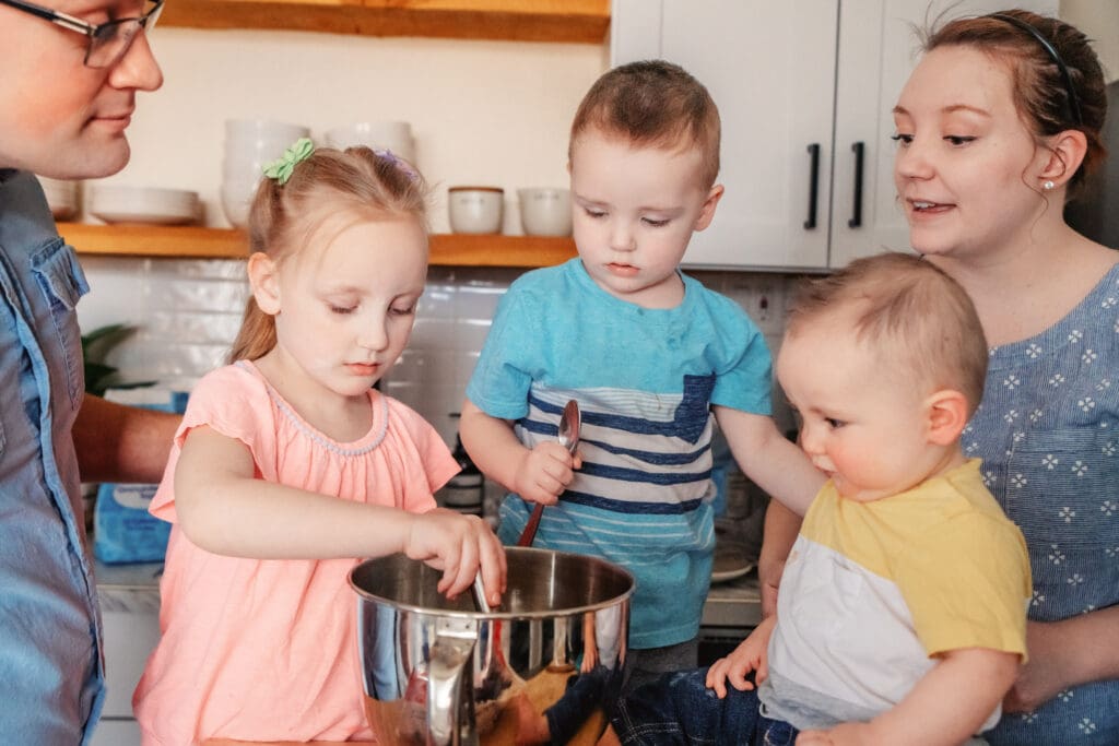 Family of five surrounding kitchen island and watching as young girl adds ingredients to mixing bowl and young boy holds spatula.
