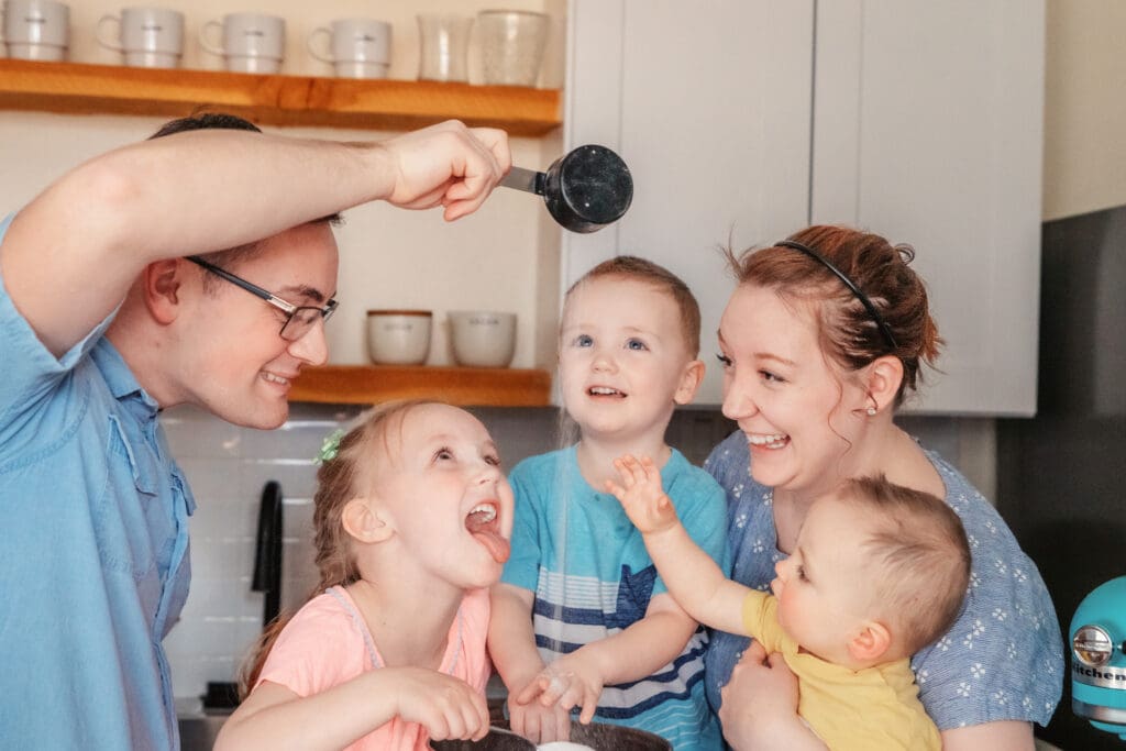 Family of five smiling and laughing as dad pours sugar from cup into mixing bowl, young girl opens her mouth to eat it, and baby boy raises his had to grab it.