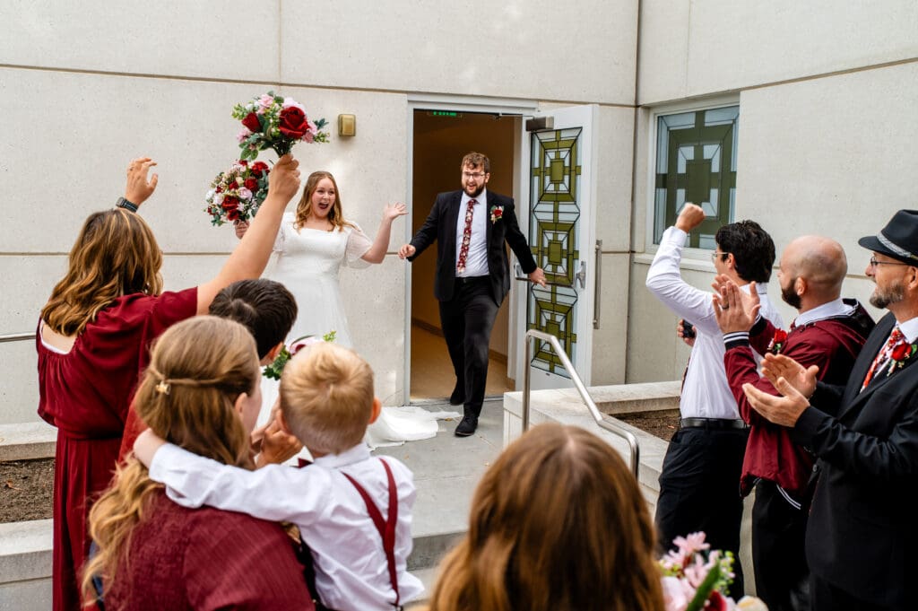 Wedding couple exits the Idaho Falls temple and friends and family cheer with bride throwing her arms in the air.