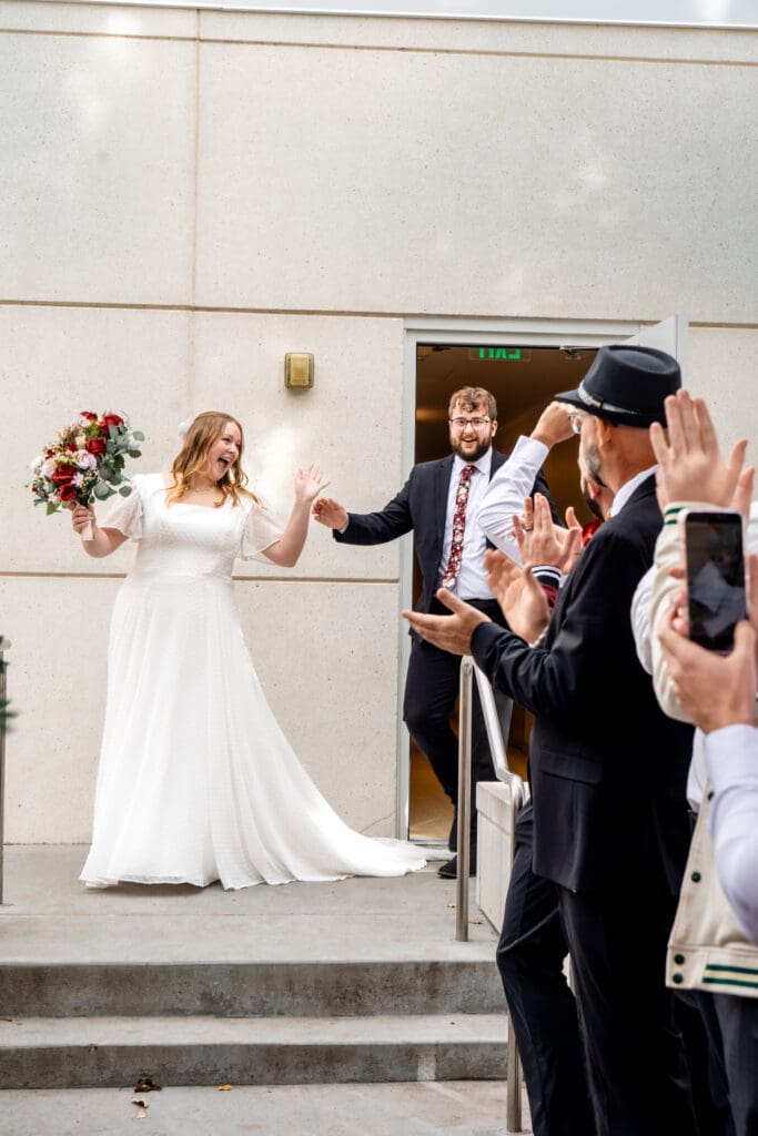 Wedding couple exits the Idaho Falls temple and friends and family clap with bride throwing her arms in the air.