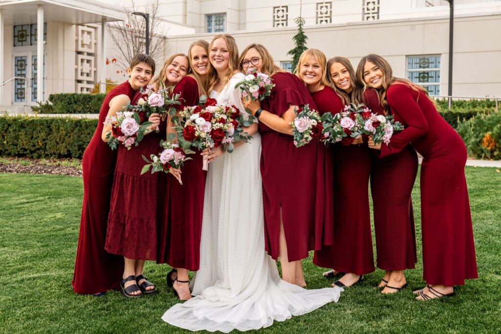 Bridesmaids and bride stand in line, hold bouquets, lean in, and smile at camera in front of Idaho Falls temple.