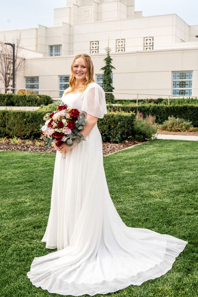 Bride holding bouquet at waist with train swirled around her as she smiles at camera in front of Idaho Falls Temple.