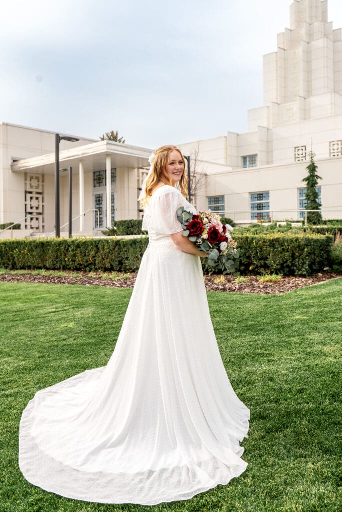 Bride looking over her shoulder and smiling at camera with her train in view in front of Idaho Falls Temple.