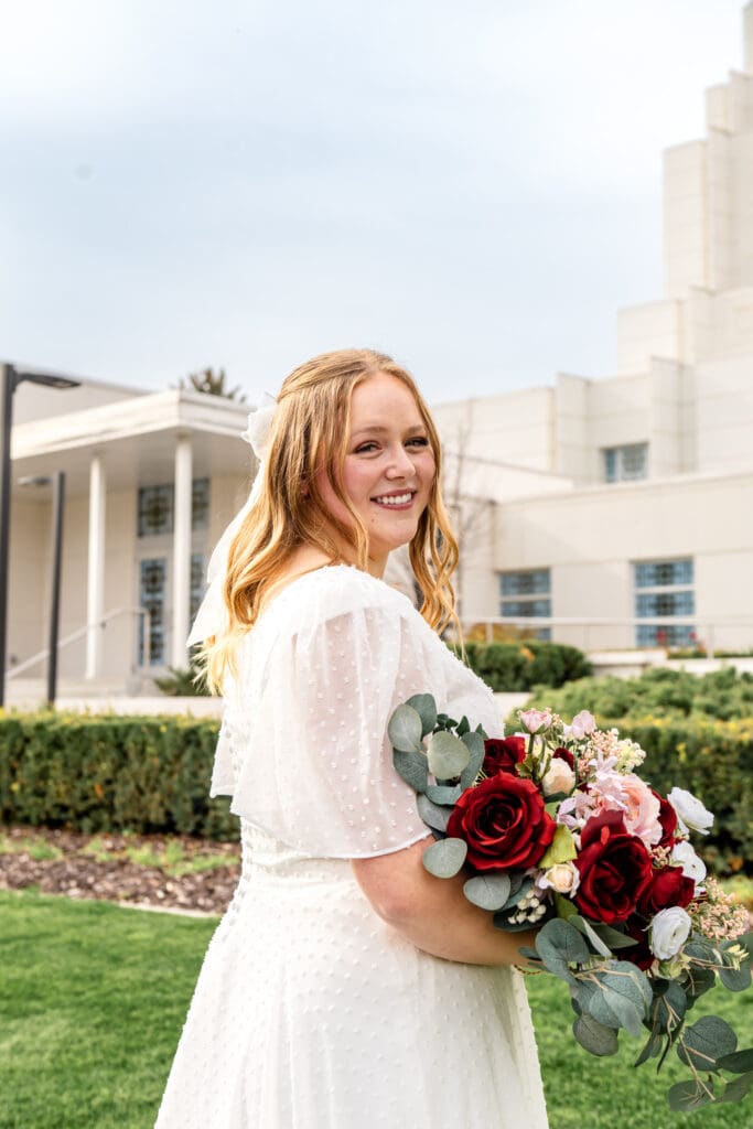 Bride looking over her shoulder and smiling at camera while hands hold bouquet at waist with beautiful blue skies and Idaho Falls Temple in the background.