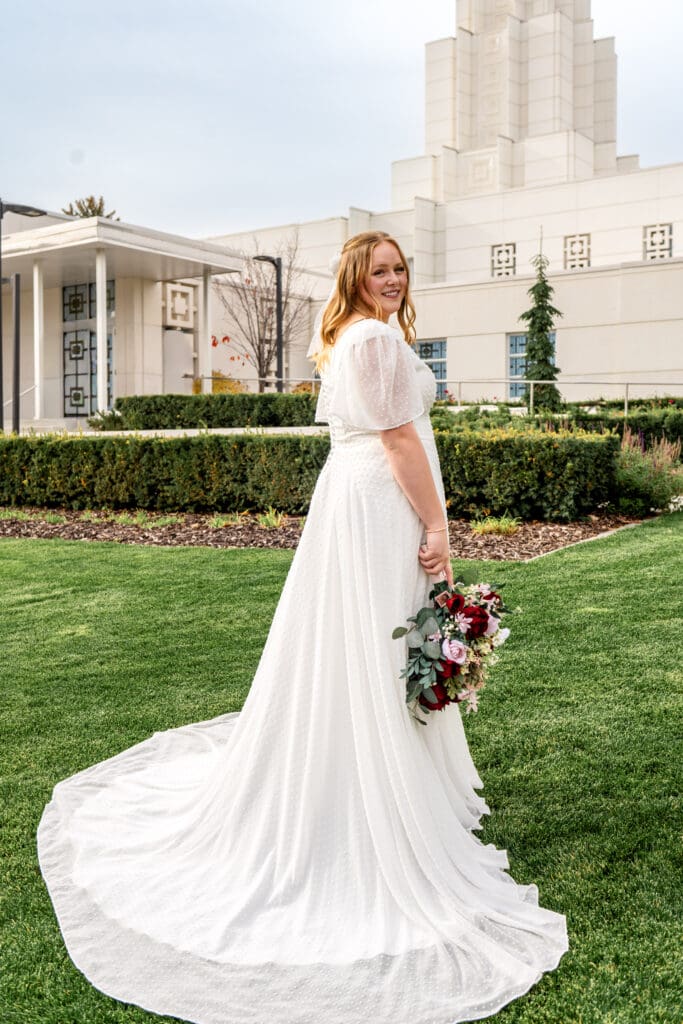 Bride looking over her shoulder and smiling at camera while right hand holds bouquet below hips with beautiful blue skies and Idaho Falls Temple in the background.
