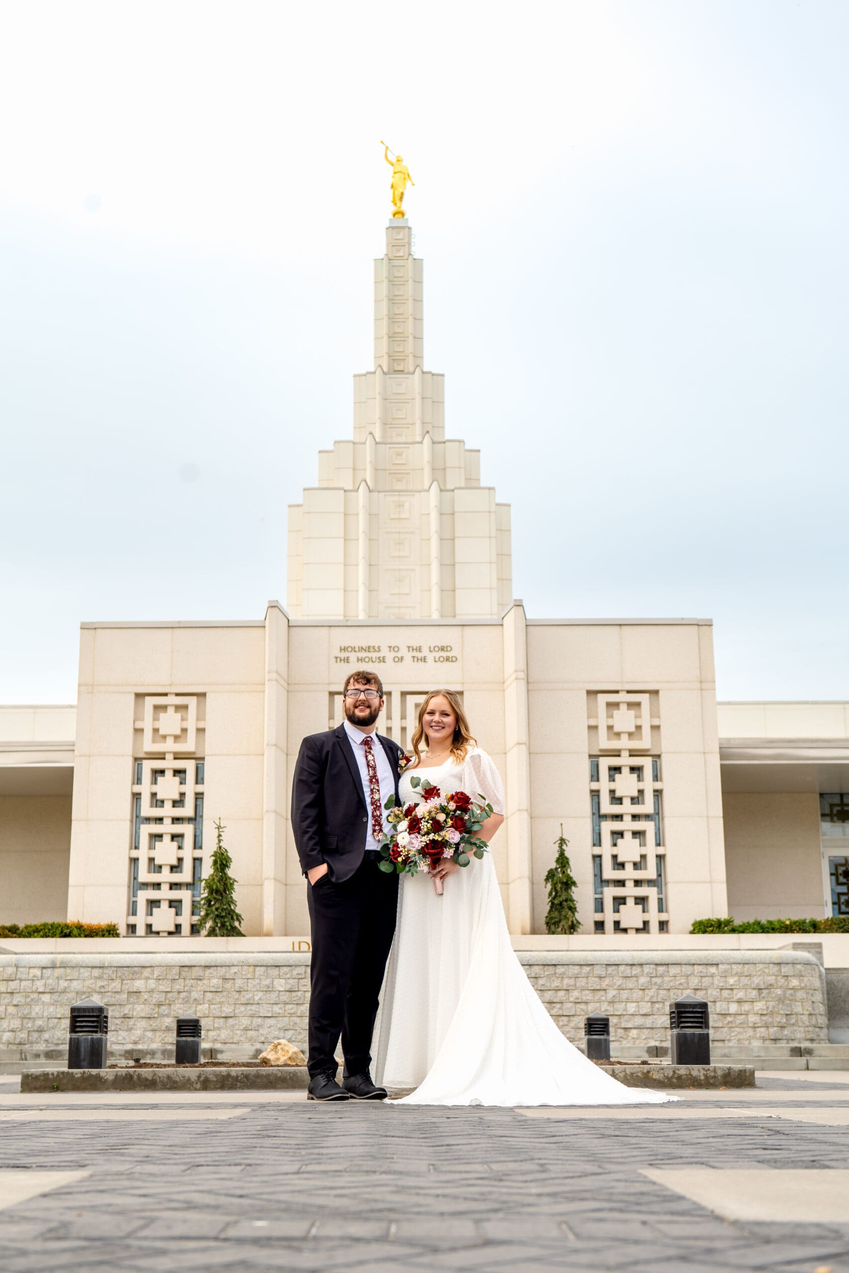 Bride and groom smiling at camera in front of Idaho Falls Temple with groom's hands in pockets and bride holding bouquet with left hand.