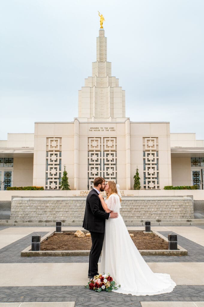 Bride and groom facing each other and kissing in front of Idaho Falls Temple with bouquet on ground.