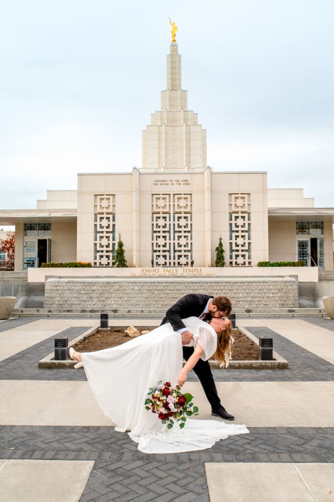 Groom dipping and kissing bride in front of Idaho Falls Temple and beautiful blue skies as bride drops bouquet down.