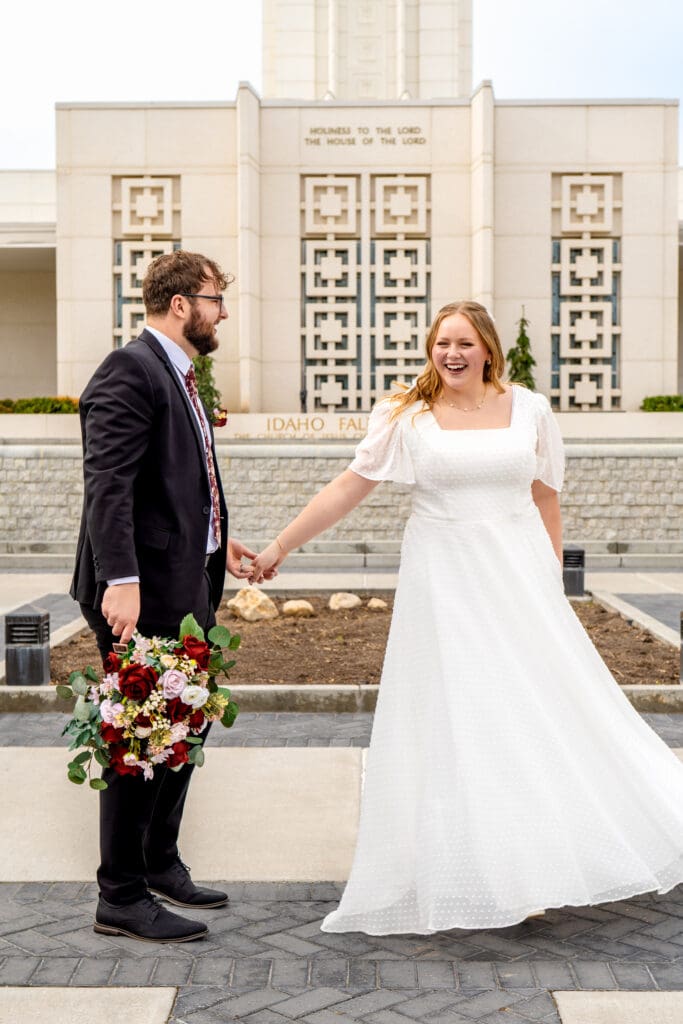 Groom holding bride's bouquet, and bride's hand as bride laughs and sways wedding dress in front of Idaho Falls Temple.
