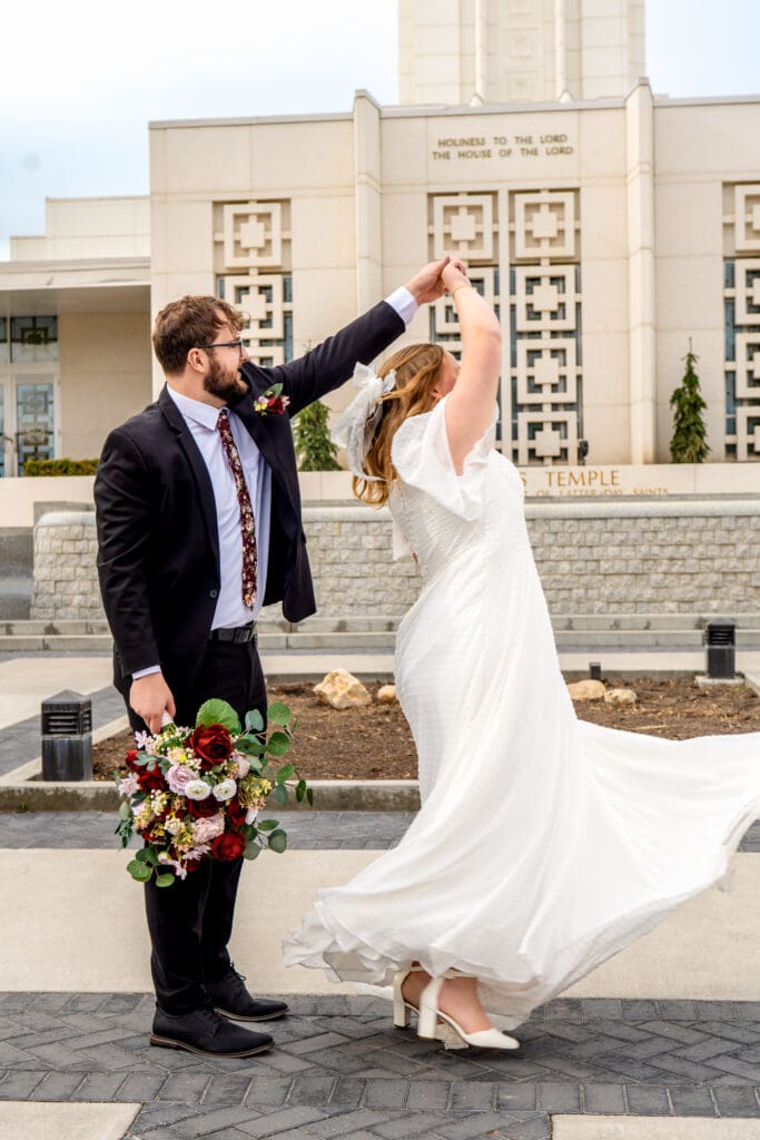 Groom holding bouquet in right hands and spinning bride with left hand in front of Idaho Falls Temple.
