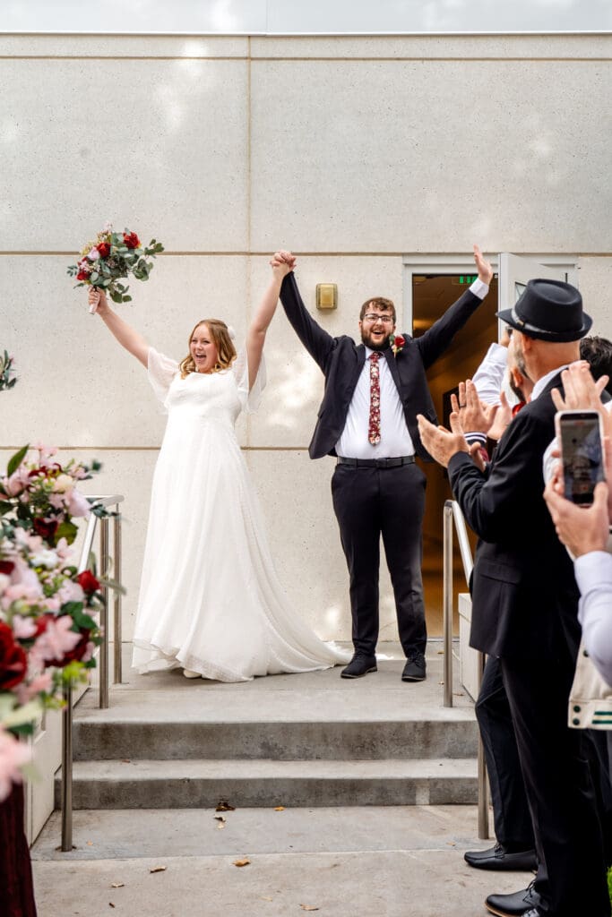 Bride and groom hold hands and throw arms in the air as friends and family clap at the Idaho Falls Temple.