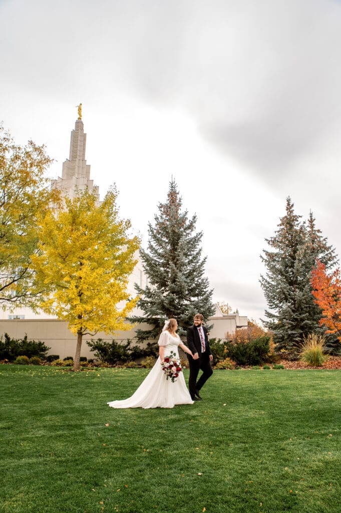 Bride and groom holding hands and talking as they walk across the grass in front of fall colored trees and the Idaho Falls temple.