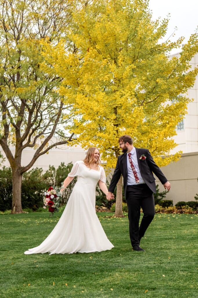 Bride and groom holding hands and laughing with groom looking back to bride as they walk across the grass in front of fall colored trees and the Idaho Falls temple.