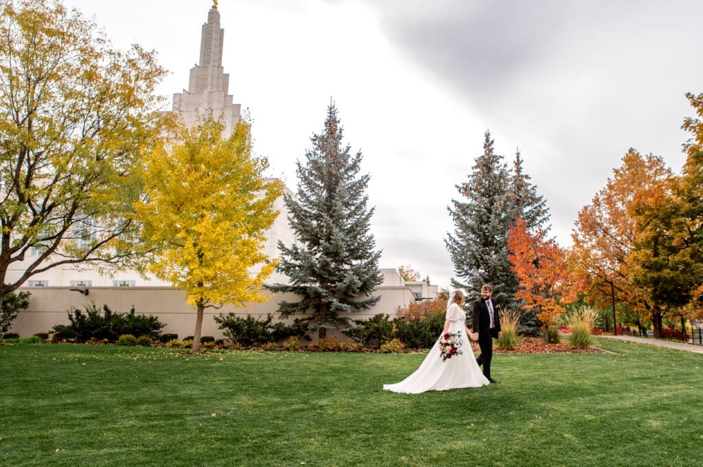 Bride and groom holding hands and smiling as they walk across the grass in front of fall colored trees and the Idaho Falls temple.
