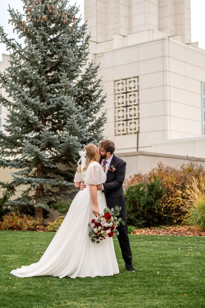 Groom kissing bride's cheek as bride holds bouquet in right hand and looks at temple on grass in front of Idaho Falls Temple.