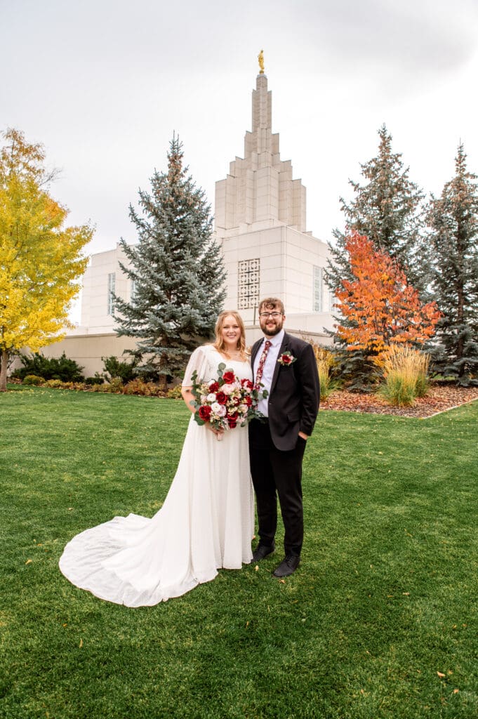 Bride and groom smiling at camera on grass in front of Idaho Falls Temple with groom's hands in pockets and bride holding bouquet with right hand.