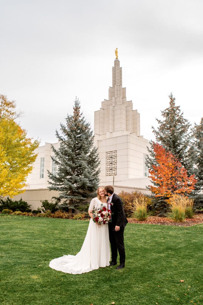 Groom kissing bride's cheek as bride holds bouquet and smiles at camera on grass in front of Idaho Falls Temple.