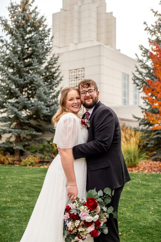Bride and groom face each other and smile at camera, touching cheeks together as bride holds bouquet below hips and groom rests his hands on bride's waist in front of Idaho Falls Temple.