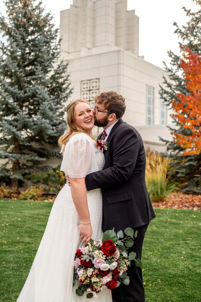 Groom kissing bride's cheek as bride holds bouquet in right hand and smiles at camera on grass in front of Idaho Falls Temple.