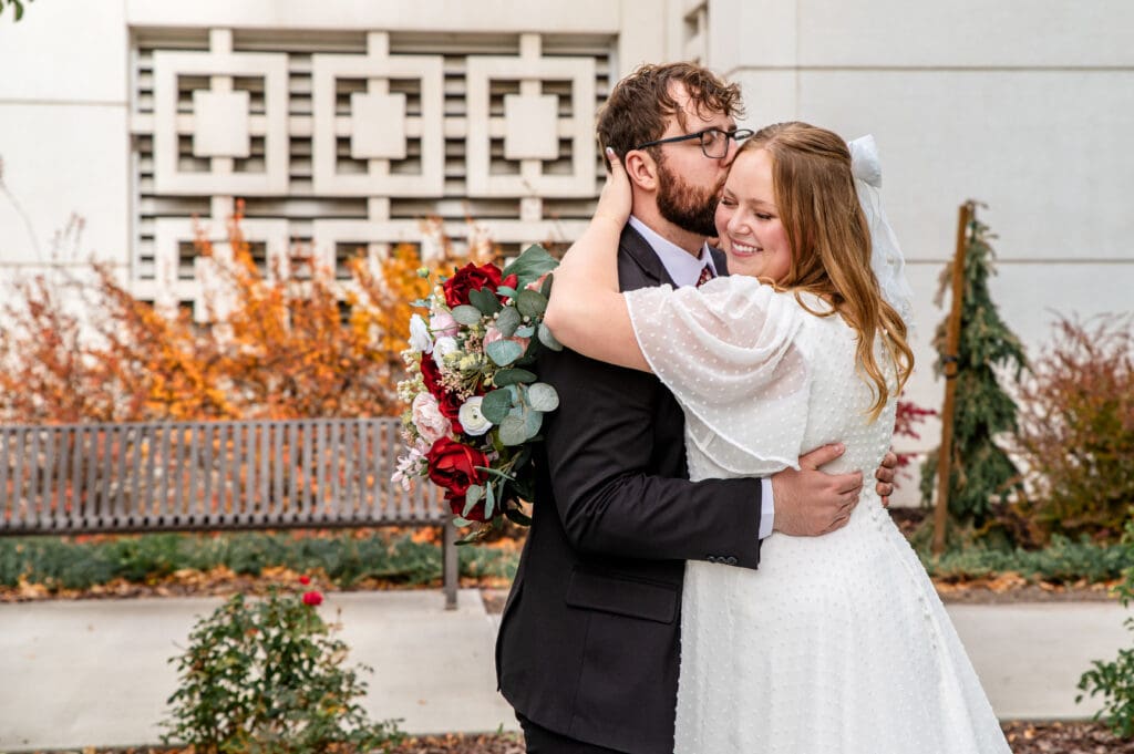 Groom holding bride and kissing her forehead as bride cups her left hand around his neck and right hands holds bouquet and she smiles with eyes closed at Idaho Falls Temple.