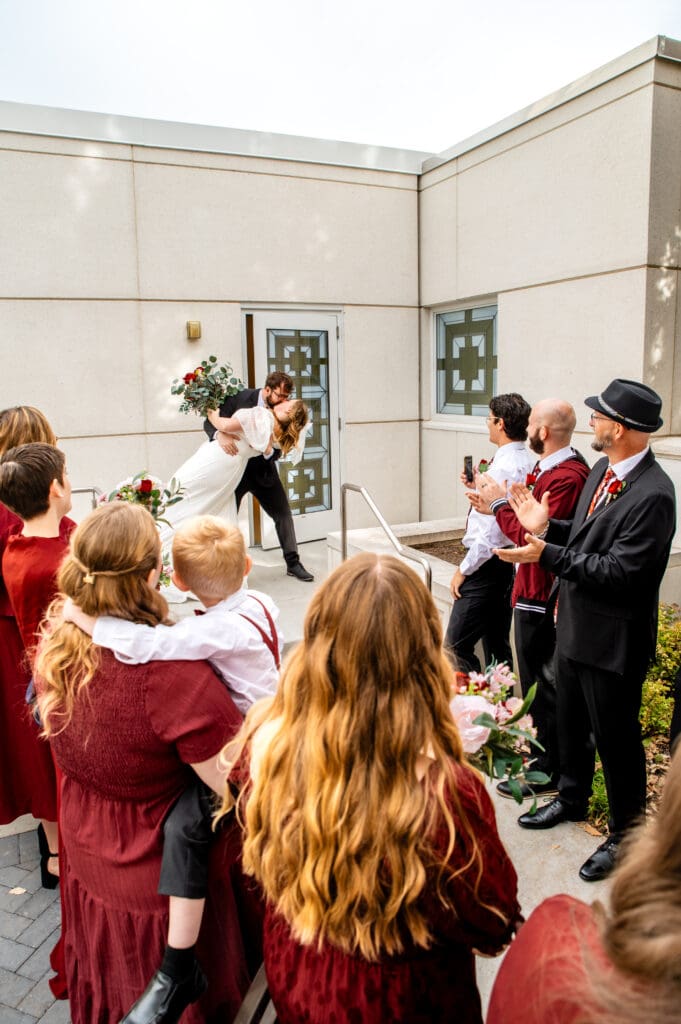 Groom dip and kissing bride on the steps of the Idaho Falls Temple bridal exit as friends and family clap.