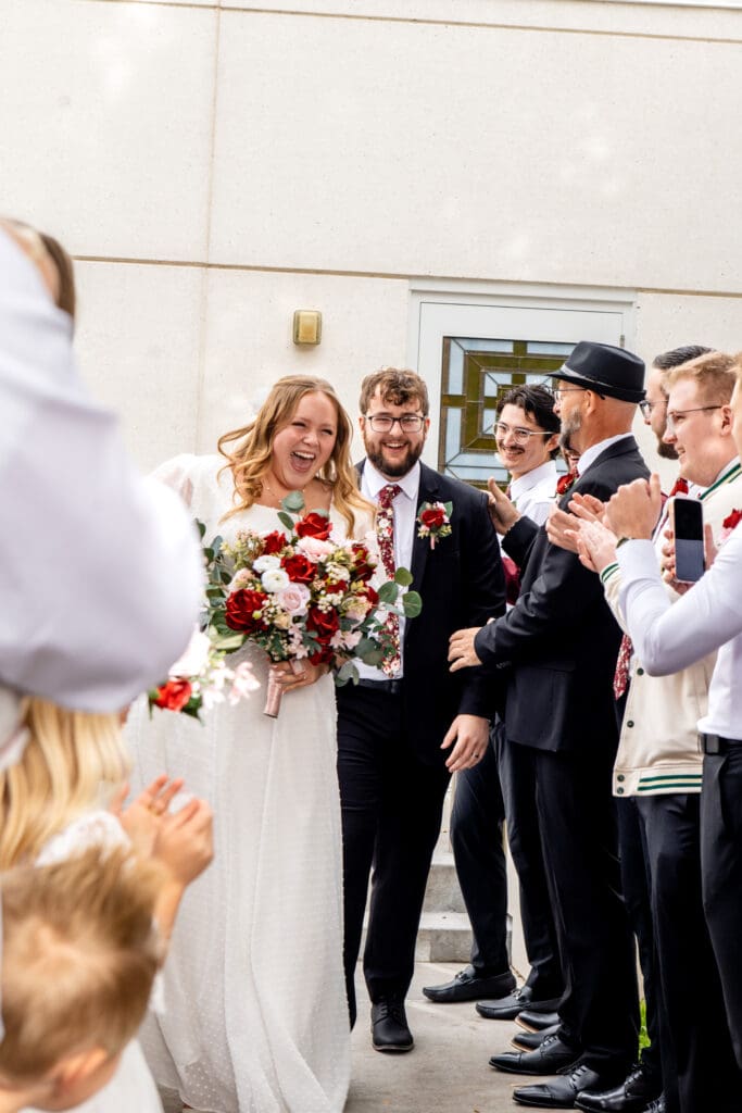 Bride and groom smile as they walk through path of friends and family clapping at Idaho Falls Temple, with the bride leading.